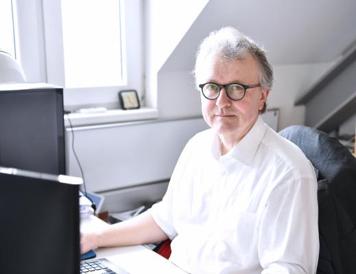 Photo of Christian Berger sitting at his desk. He wears a white shirt and glasses with a black and brown frame. He looks straight into the camera.