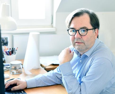 photo of Gert M.Mayr-Keber sitting at his desk, looking into the camera. He is wearing a blue shirt and black glasses.