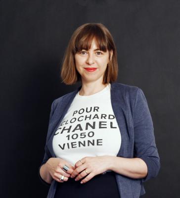 Photo of Johanna Mayr-Keber in front of a dark background. She wears a tshirt with the words "pour chlochard chanel 1050 vienne", which is by the artist Martin Grandits.